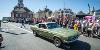 An old Ford Mustang, green in colour, travels along Stockton High Street. Crowds line the route. Stockton Town Hall is visible in the background, and you can just make out some other supercars following.