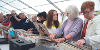 A row of people browsing through rows of records at a Record Fair - the focal point of the image is a girl with brown hair in yellow jumper holding up a Tracy Chapman record while talking to an older lady with white hair and wearing a lilac cardigan.